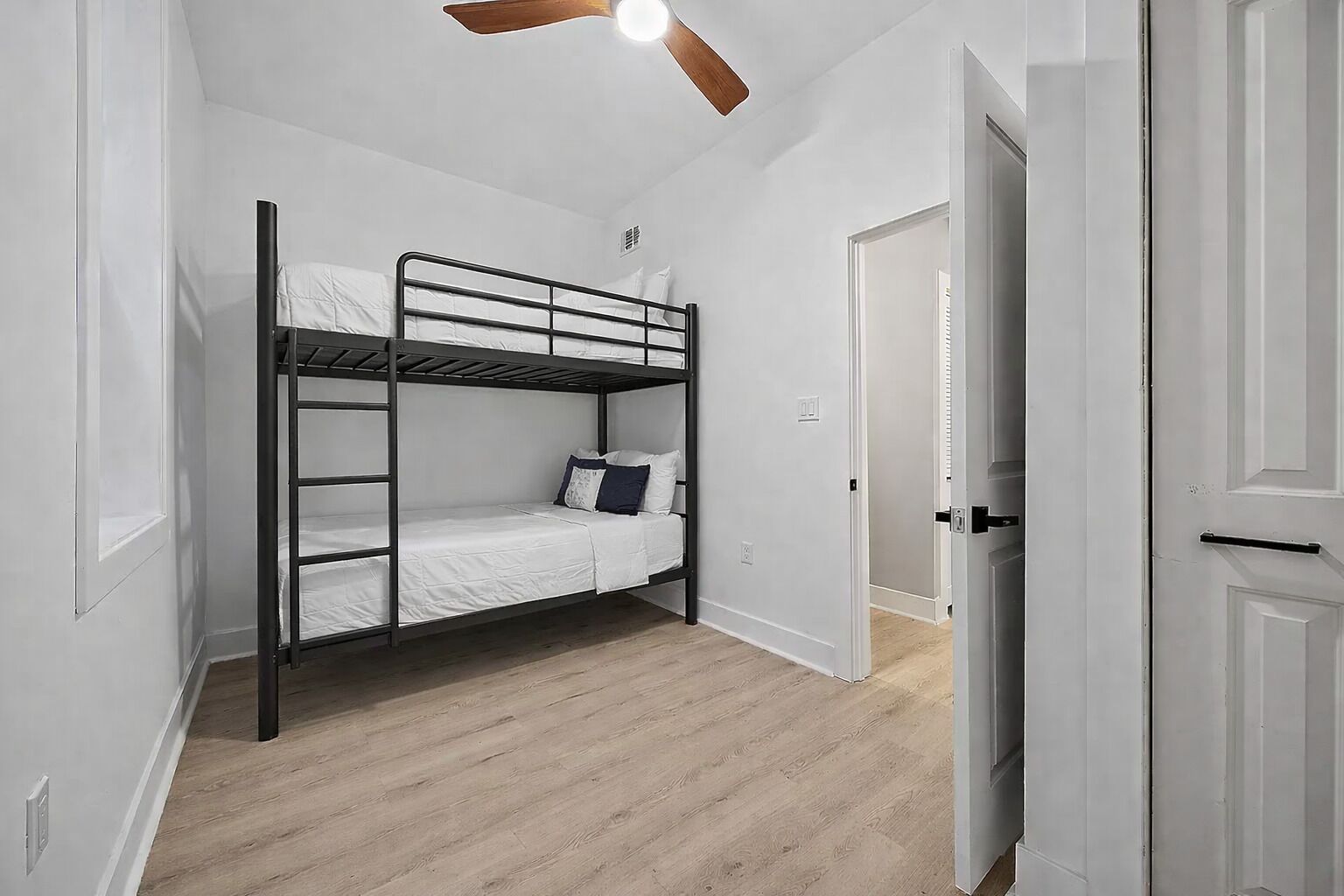 A bunk bed room with a ceiling fan, white walls, and wooden flooring.