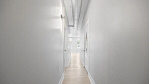 A long, narrow hallway with white walls and a wooden floor, leading to a door at the end.