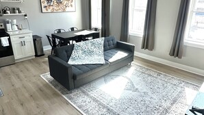 A modern living room with a grey sofa, a dining table, and a kitchen area in the background.