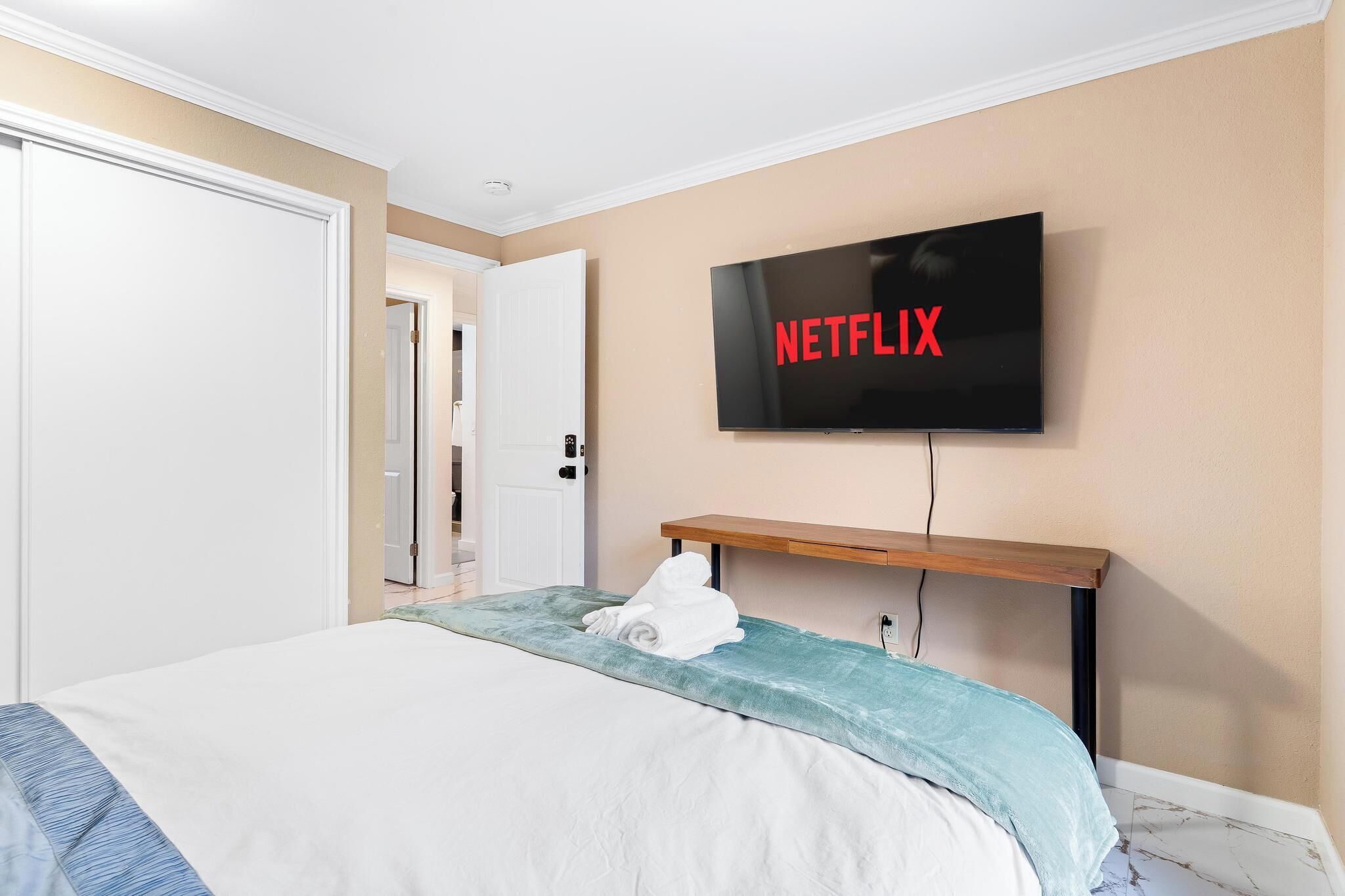 Habitación de hotel con una cama, un televisor sujeto a la pared mostrando el logotipo de Netflix, y un estante de madera debajo del televisor.