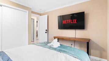 Habitación de hotel con una cama, un televisor sujeto a la pared mostrando el logotipo de Netflix, y un estante de madera debajo del televisor.