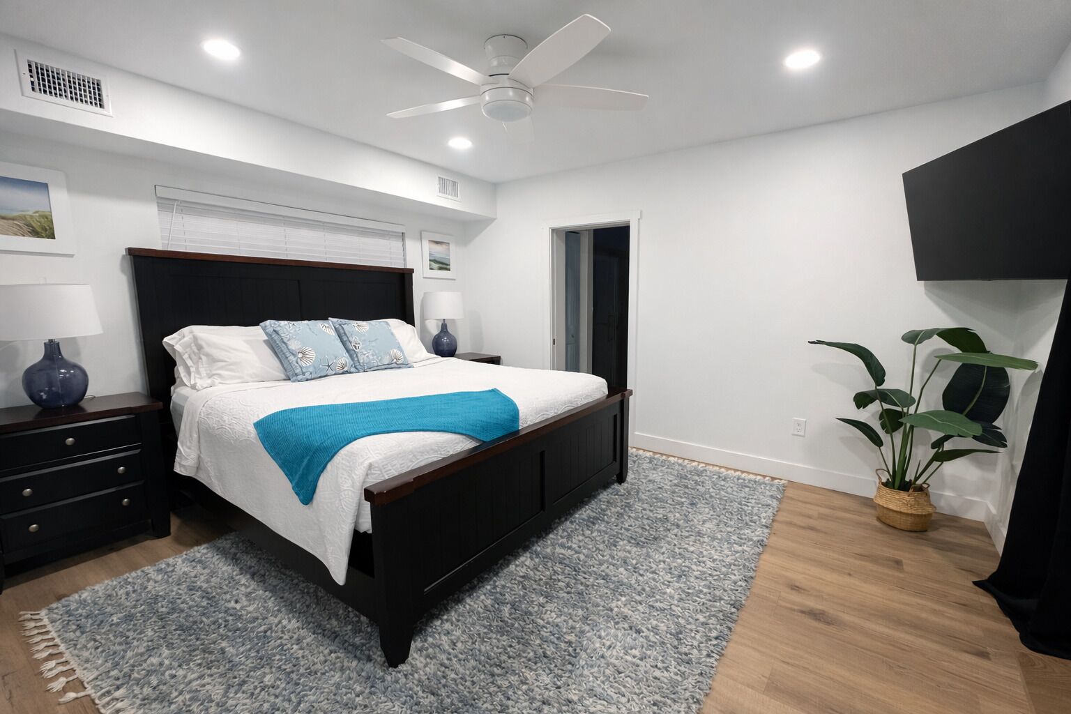 A bedroom with a bed, bedside tables, a ceiling fan, a potted plant, and a door.
