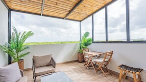 Eine überdachte Terrasse mit Holzboden, einem Tisch, Stühlen, einem kleinen Hocker und zwei Blumentöpfen.