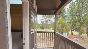 Una terraza de madera con barandilla, un dormitorio con cama y ventana, y vistas a árboles y césped.