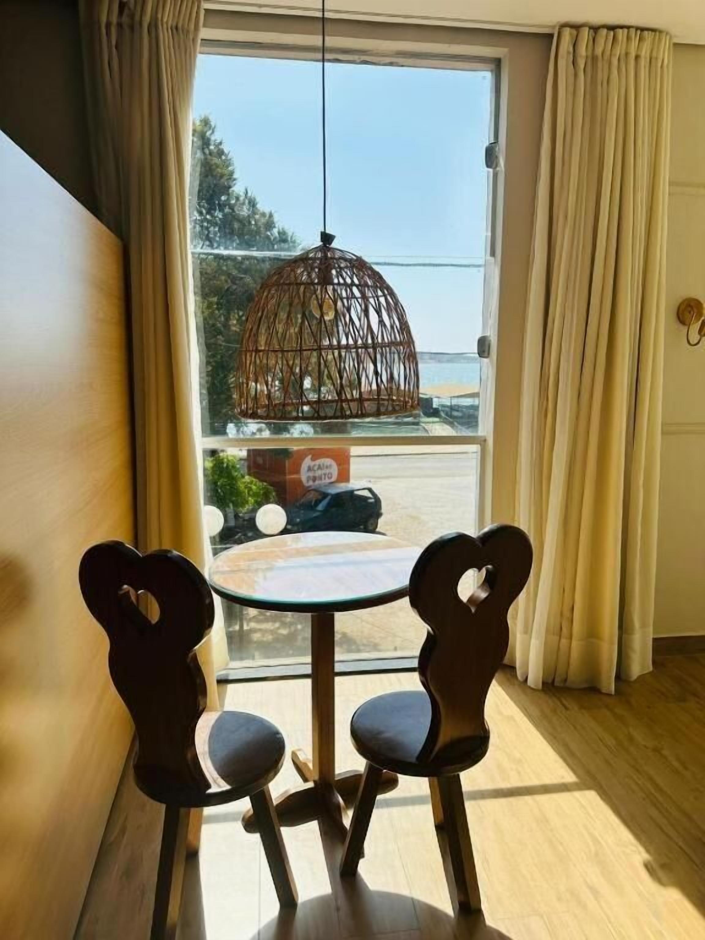 Una habitación con dos sillas con forma de corazón, una mesa redonda, un lámpara colgante y vista a un estacionamiento a través de un ventanal grande.