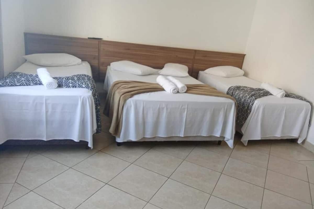 Een hotelkamer met drie enkele bedden, elk met een witte spreidek en een opgerolde handdoek. De kamer heeft een betegelde vloer en een houten hoofdbord.