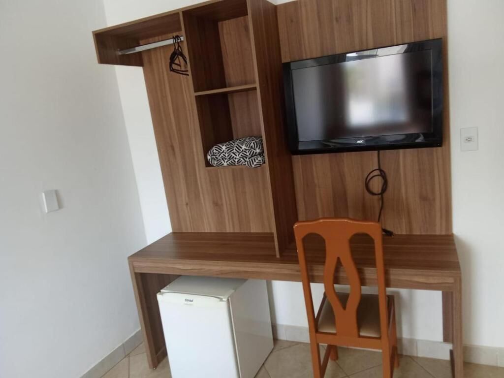 A compact living space with a wooden desk, a television mounted on the wall, a small refrigerator, and a chair.