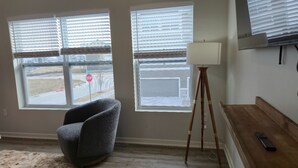 Ein Zimmer mit zwei Fenstern, einem Sessel, einer Stehlampe und einem an der Wand befestigten Fernseher.