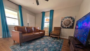 Un salón con un sofá de cuero marrón, un tapete azul con estampado, una mesa de centro de madera, un mueble de televisión de madera y un televisor grande que muestra un paisaje urbano.