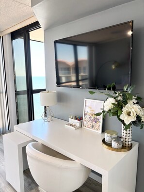 A modern room with a large flat-screen TV mounted on the wall, a white desk with a lamp, a framed picture, and a vase with flowers. - 20th-Floor Oceanfront Condo with Balcony, Pools, Beach Access & Resort Amenities (Ocean City)