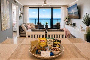 A modern living room with a large window overlooking the sea, a dining area with a tray of snacks, and a cozy seating arrangement. - 20th-Floor Oceanfront Condo with Balcony, Pools, Beach Access & Resort Amenities (Ocean City)