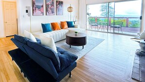 A spacious living room with a dark blue sofa, a white sofa with colorful cushions, and a round coffee table. There are three framed pictures on the wall, a floor lamp, and a sliding glass door leading to a balcony with outdoor furniture.