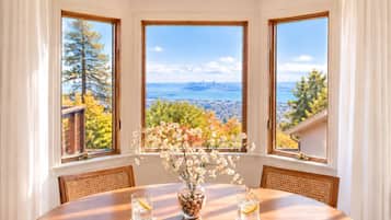 Un coin repas comprenant une table, des chaises, un vase avec des fleurs et une vue sur le paysage urbain, visible à travers trois fenêtres.