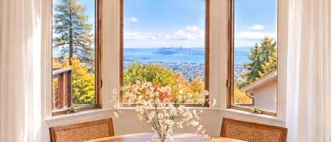 A dining area with a table, chairs, a vase with flowers, and a view of a cityscape through three windows.