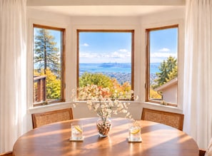 A dining area with a table, chairs, a vase with flowers, and a view of a cityscape through three windows.