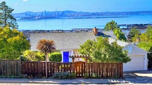 A residential area with a house featuring a blue door, a wooden fence, and a view of a city skyline across a body of water.