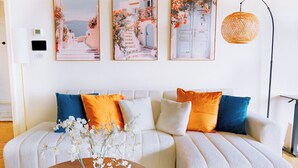 A living room with a white sofa, orange and blue cushions, a wooden coffee table with a vase of white flowers, and three framed pictures on the wall.