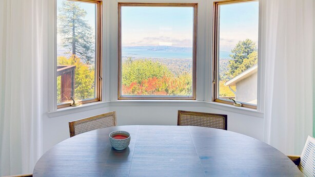 A dining area with a table and chairs, three windows with a view of trees and a bridge, and a bowl of red liquid on the table.