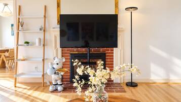 A living room with a fireplace, a flat-screen TV mounted above it, a wooden floor, a round coffee table with a vase of flowers, a teddy bear decoration, and a bookshelf with various items.