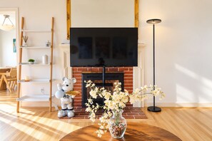 A living room with a fireplace, a flat-screen TV mounted above it, a wooden floor, a round coffee table with a vase of flowers, a teddy bear decoration, and a bookshelf with various items. - Cozy Berkeley Hills Home with Stunning Views (Berkeley)