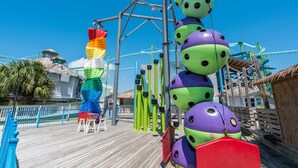 A wooden deck with a thatched-roof building, a colorful play structure, and a water slide. - Boardwalk A18 | Beach Access & Pool | New Listing! (Pensacola Beach)