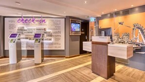 Un espace d’accueil d’hôtel comprenant un comptoir d’enregistrement, un écran numérique et un mur présentant divers équipements et services.