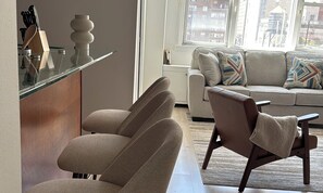 A modern living room with a sofa, armchairs, a glass coffee table, and a kitchen area with utensils.