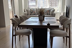A dining area with a rectangular table, six chairs, a vase with dried plants, and a sofa with patterned cushions in the background.