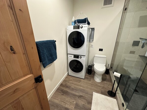 Un baño pequeño con puerta de madera, lavadora, inodoro y cabina de ducha de vidrio.