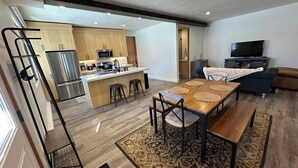 Eine moderne Küche mit Edelstahlgeräten, eine Essecke mit Holztisch und schwarzen Stühlen sowie ein Wohnzimmer mit Sofa und Fernseher.