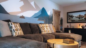 Ein modernes Wohnzimmer mit einem Ecksofa, einem runden Couchtisch und einer Wandmalerei von Bergen und einer Burg.