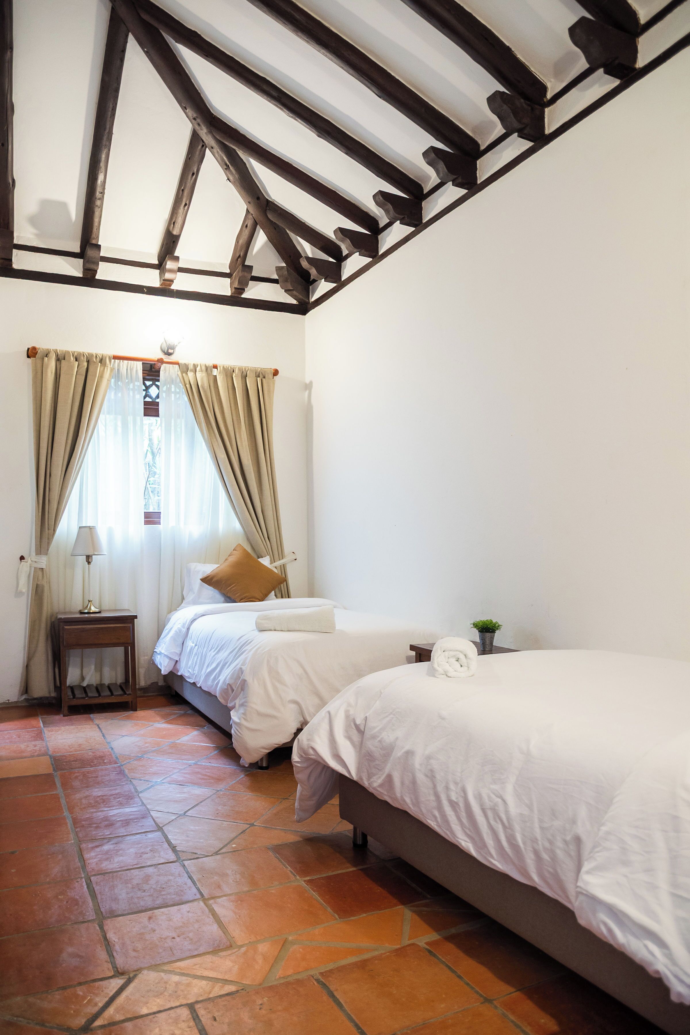 Un dormitorio con una cama, una mesita de noche, una lámpara, una ventana con cortinas y suelo de baldosas.