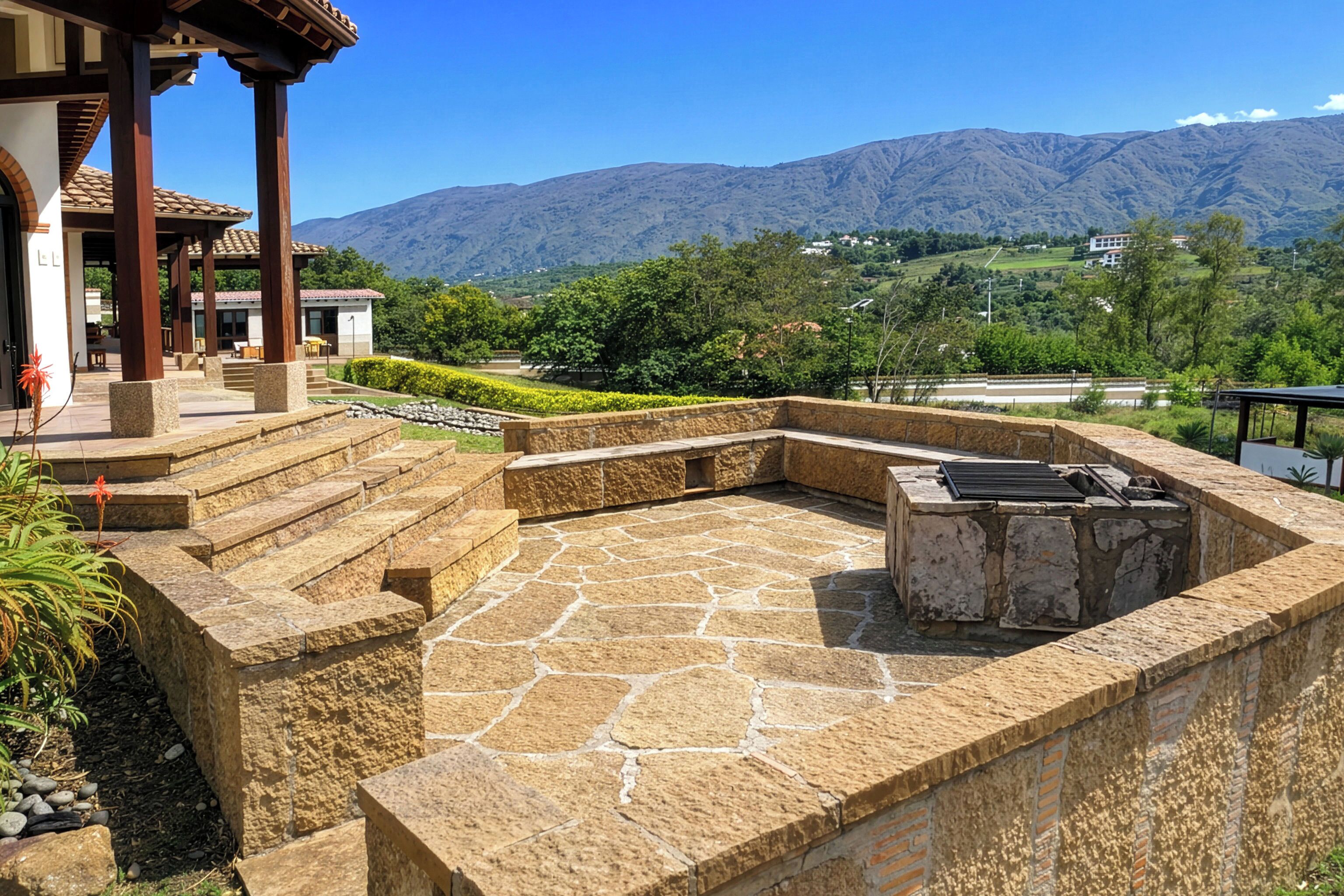 Un patio de piedra con una parrilla, escalones que llevan a una zona cubierta y montañas al fondo.