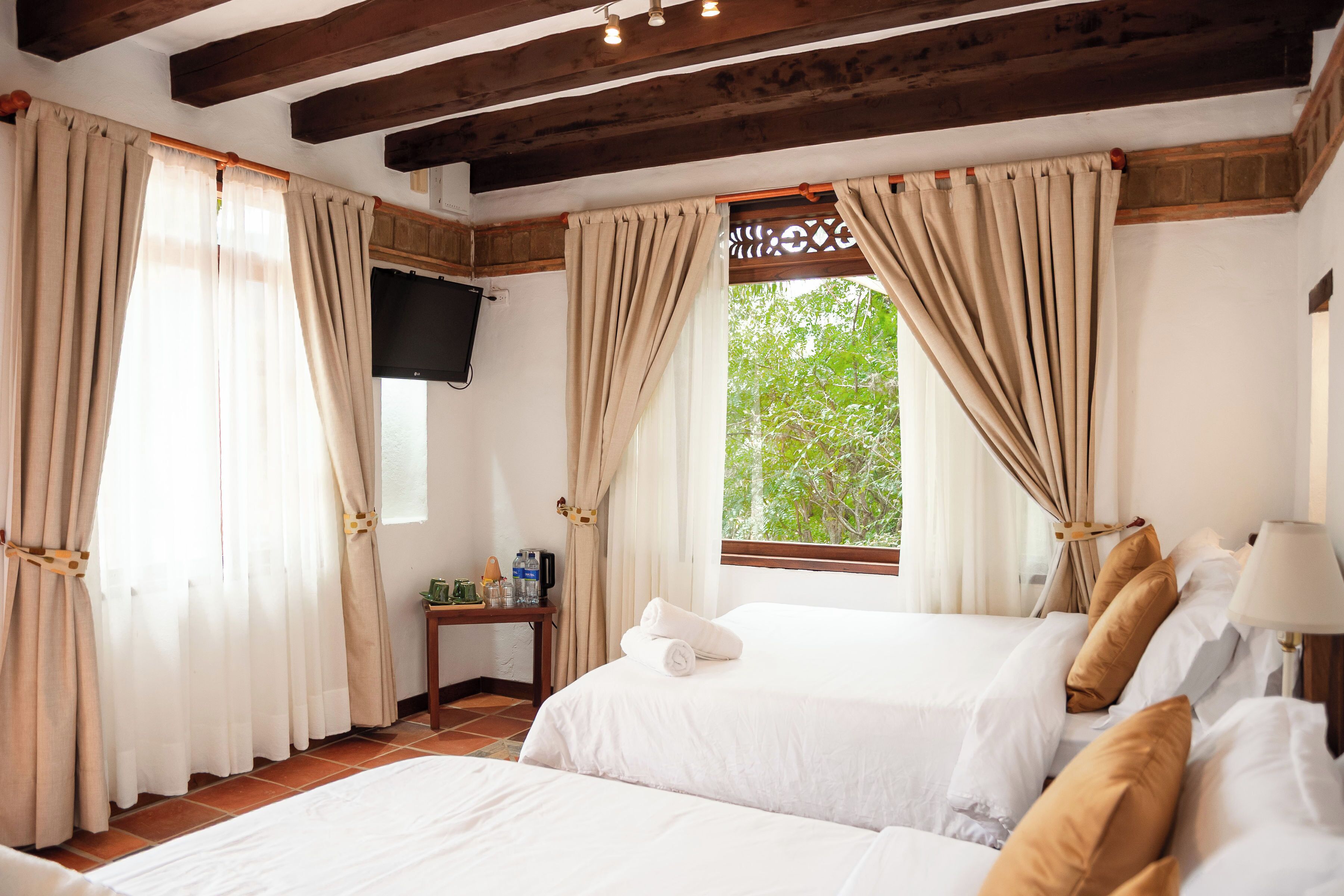 Habitación de hotel con dos camas, cortinas color beige, un televisor en la pared y una ventana con vistas a zonas verdes.
