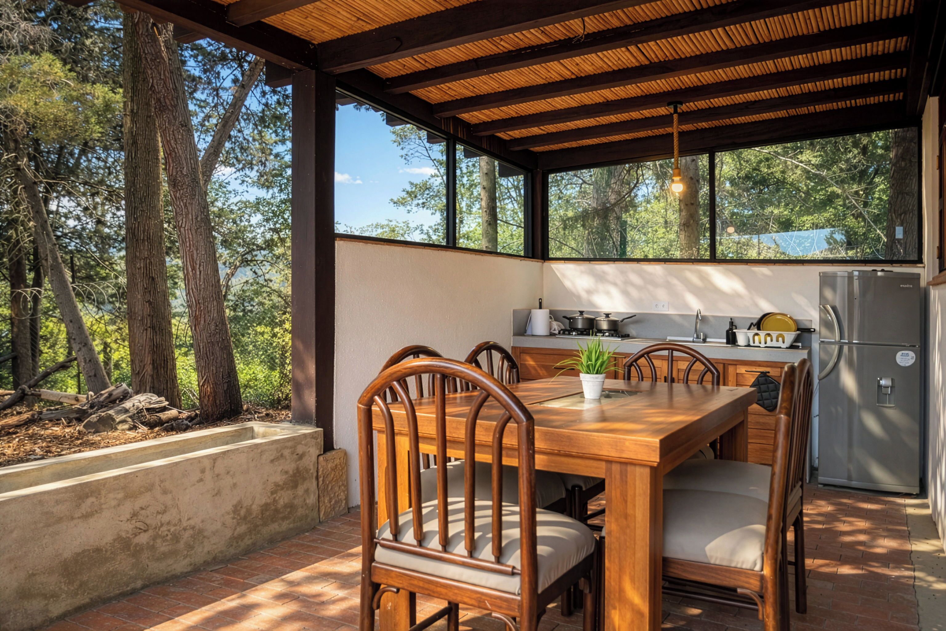 A covered outdoor dining area with a wooden table and chairs, a potted plant, a kitchen area with a refrigerator, and a view of trees.