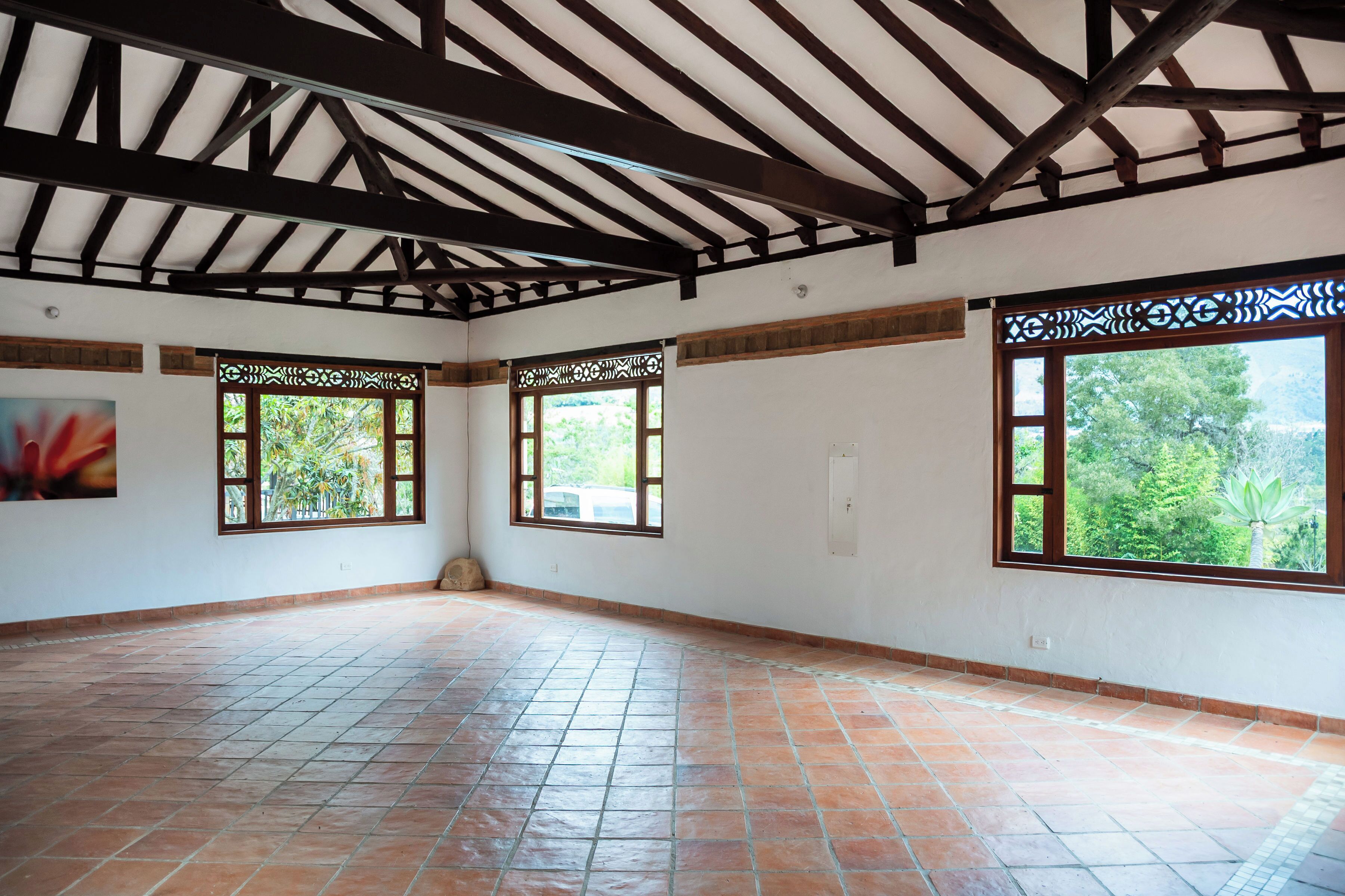 A spacious room with a tiled floor, wooden ceiling beams, and large windows with decorative frames.
