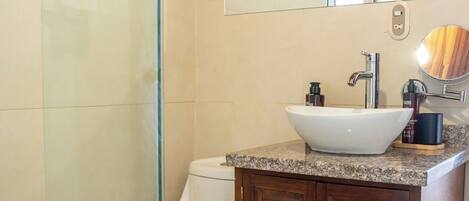 Un baño con cabina de ducha de vidrio, un lavabo blanco sobre un mueble de madera y un espejo grande sobre el mueble.