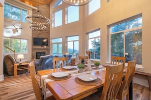 A spacious dining area with a wooden table set for four, surrounded by wooden chairs. A stone fireplace and a large TV are visible in the background. - Upscale Mountain Retreat, Hot Tub & Game Room! (Helen)