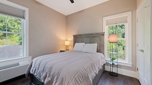 A bedroom with a bed, a ceiling fan, a heater, a window with blinds, and a nightstand with a lamp. - Brand New Home- Maggie Valley Sunset (Waynesville)