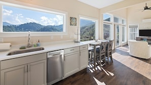 A modern kitchen with large windows, a dishwasher, and a dining area with a view of the mountains. - Brand New Home- Maggie Valley Sunset (Waynesville)