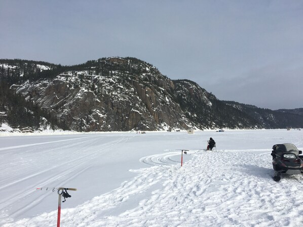 Un paysage enneigé avec une montagne en arrière-plan, une personne qui fait du ski et un véhicule garé dans la neige.