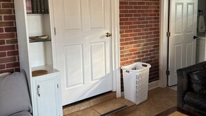 A living room with a white door, a brick wall, a grey couch, a wooden cabinet, and a wicker basket.