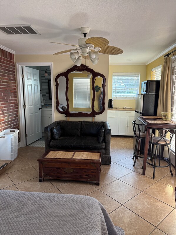 A living room with a dark leather sofa, a wooden coffee table, a ceiling fan, and a kitchen area with a refrigerator and microwave.
