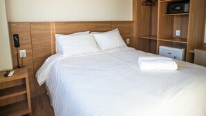 Una camera d'albergo con una testiera in legno, un letto con lenzuola bianche, un comodino con telefono e telecomando, e un piccolo frigorifero.