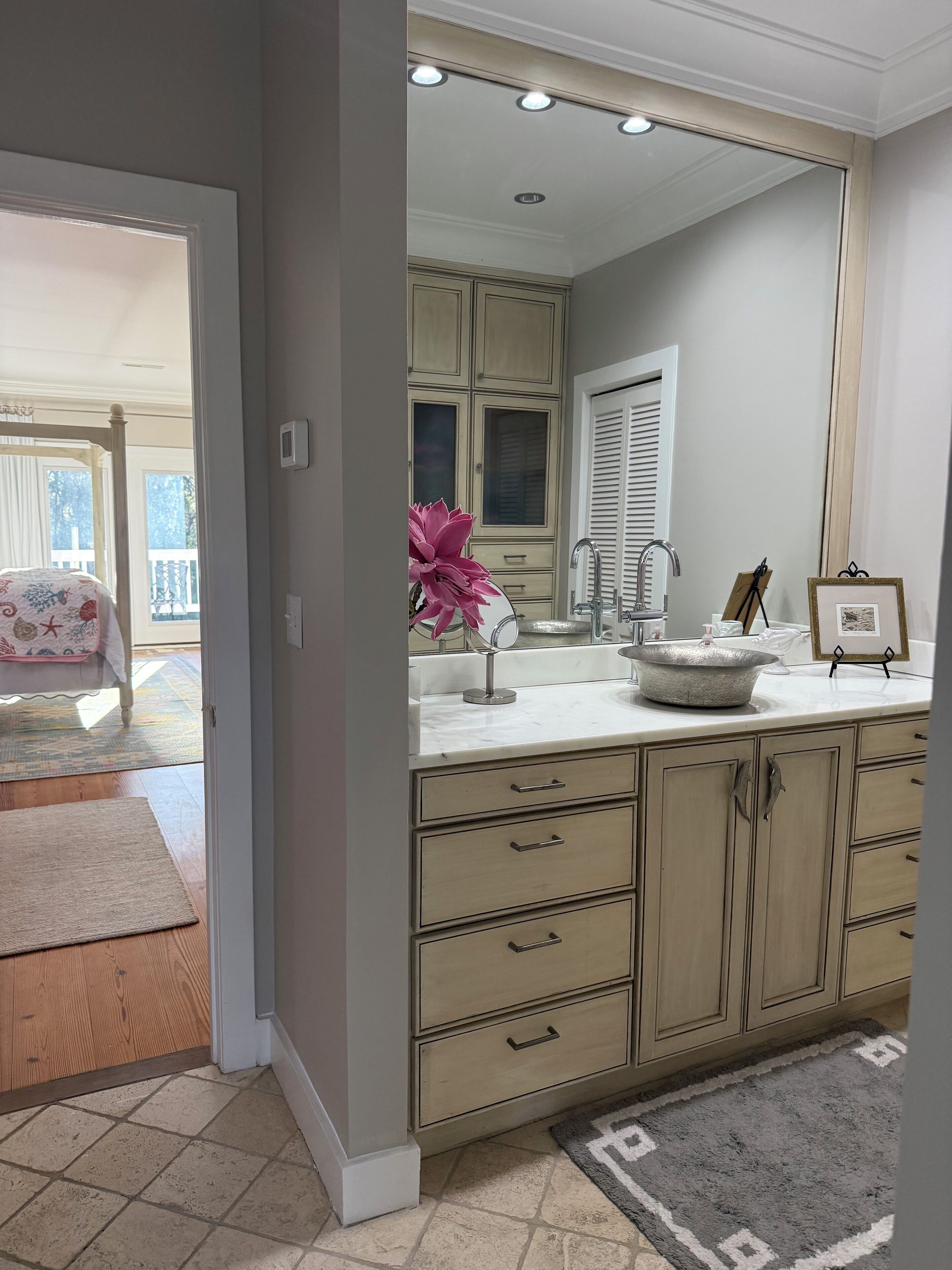A bathroom with a large mirror, a sink, and a vanity with drawers. There is a window with blinds and a door leading to another room.