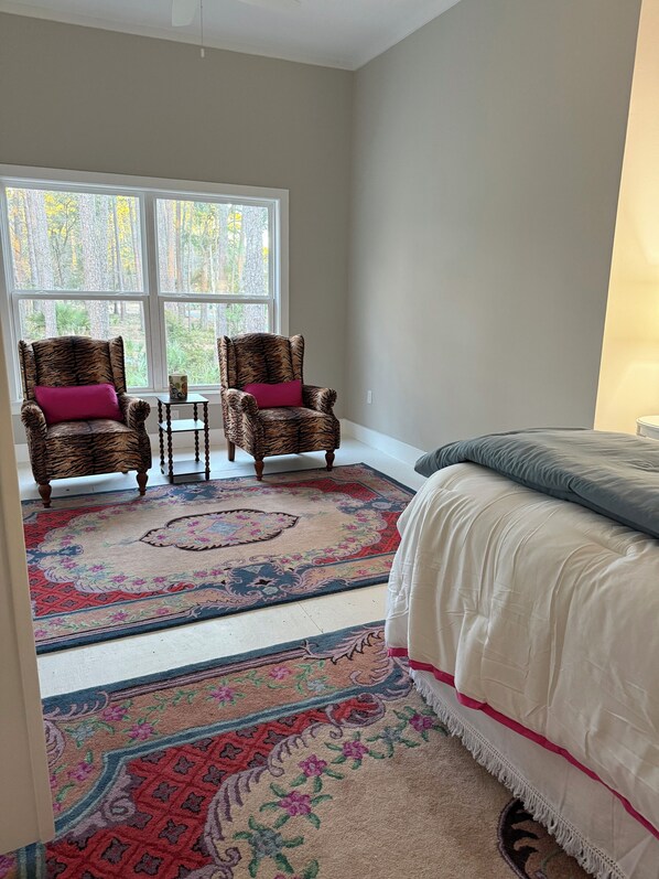 A bedroom with a bed, two tiger-patterned chairs, a small table, and a large window with a view of trees.