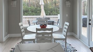 A dining area with a round table, white chairs, a chandelier, and a view of a deck and trees. - Pink Paradise aka The Purple House (Daufuskie Island)