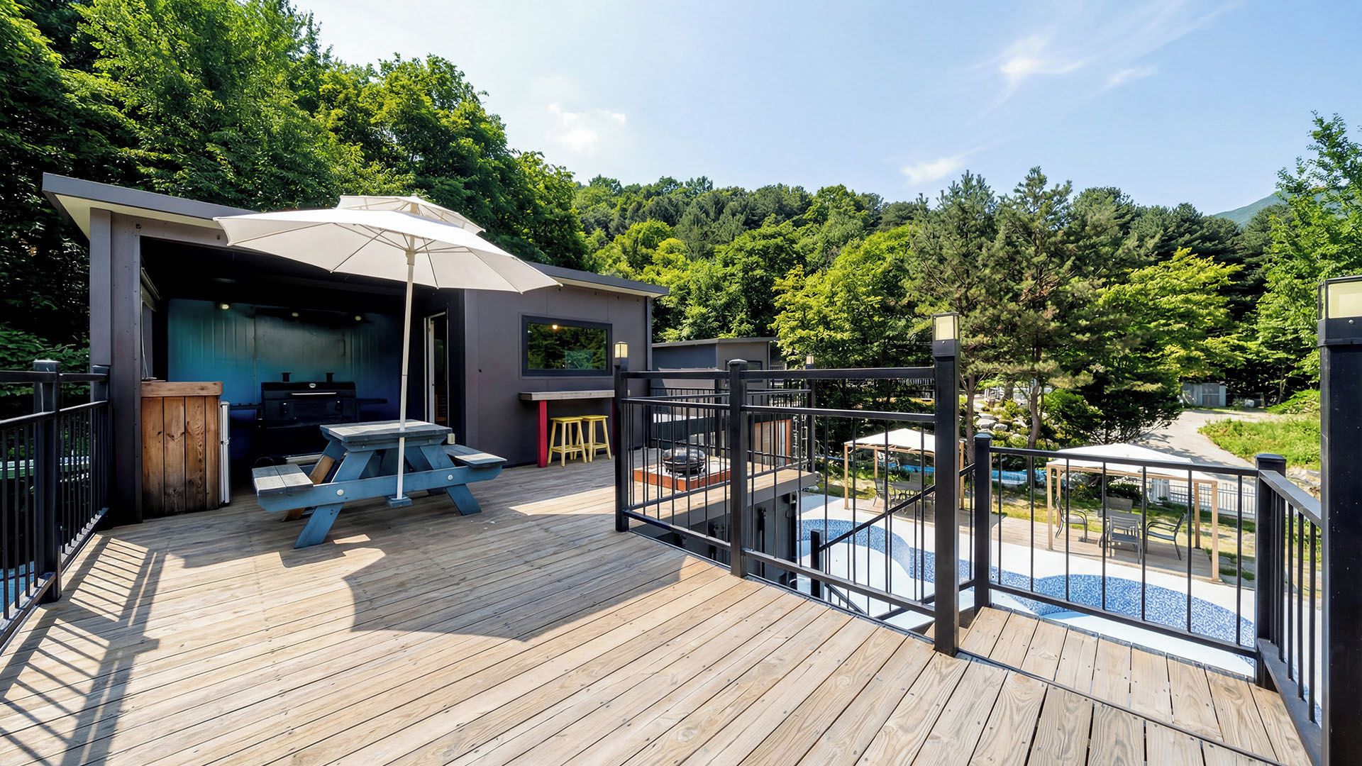 Una terraza de madera con zona de barbacoa, espacio de estar cubierto y piscina.