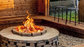 Un foyer circulaire en pierre, avec des flammes, entouré d’une terrasse en bois et d’une fenêtre donnant sur un paysage verdoyant.