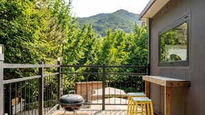 A balcony with a barbecue grill, wooden stools, and a view of a forested mountain. - Gapyeong Ruba Pension (Gapyeong)